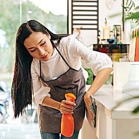 A young female professional house cleaner of Asian ethnicity wearing a white long sleeve shirt and a charcoal-colored apron uses a pale blue rag and orange spray bottle to clean a kitchen countertop in an upscale home.