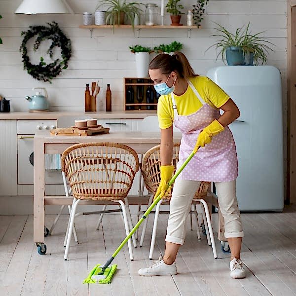 A young female professional house cleaner wearing a yellow shirt and a pink apron uses a swifer sweeper to clean a tile floor in an eclectic-style kitchen.