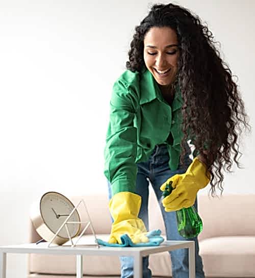 An attractive and professional young female house cleaner wearing a green collared shirt and yellow rubber cloves uses a blue rag and green spray bottle to clean a living room coffee table.