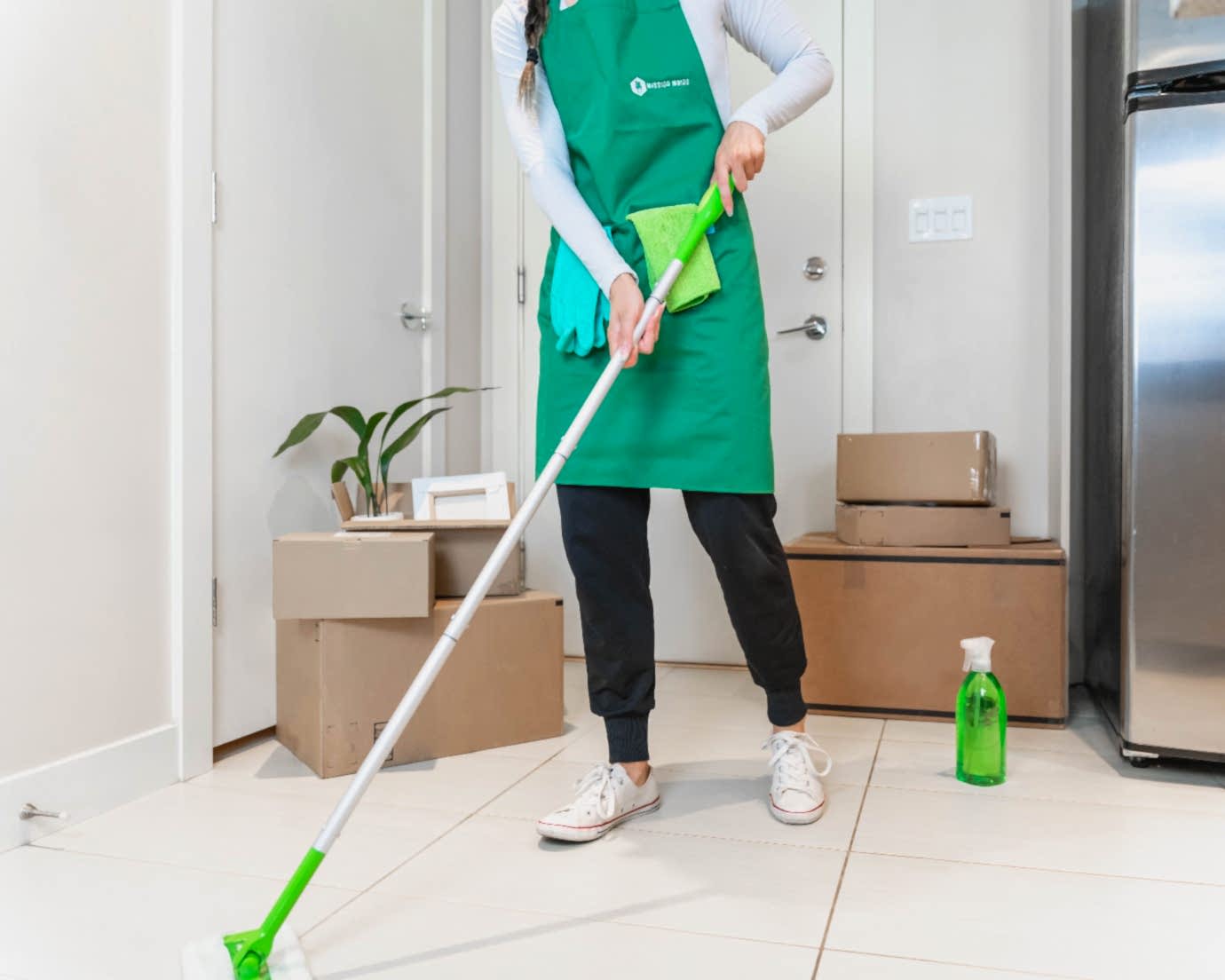 A young female professional house cleaner wearing a white long-sleeve shirt and a green apron uses a Swifer Sweeper to clean a tile floor in a Calgary condo kitchen.