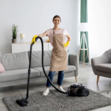 A young cleaning lady holds a vacuum while smiling into the camera.