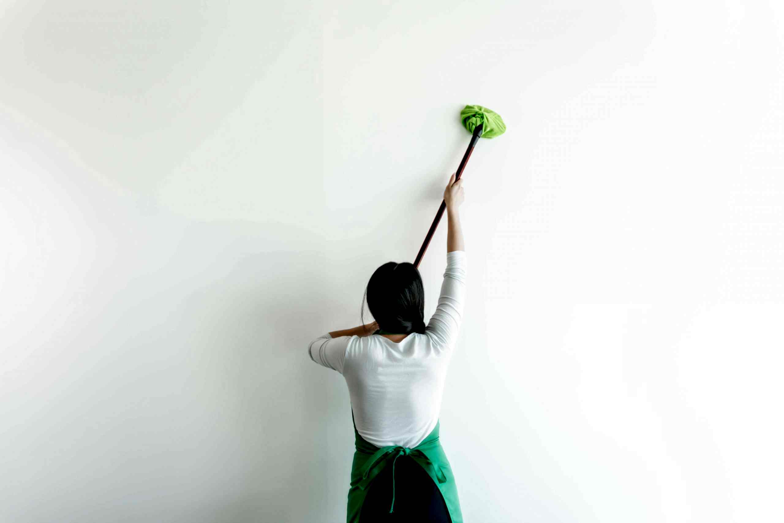 A young female professional house cleaner wearing a white long-sleeve shirt and a green apron uses a cleaning tool to clean a wall in a Calgary home.