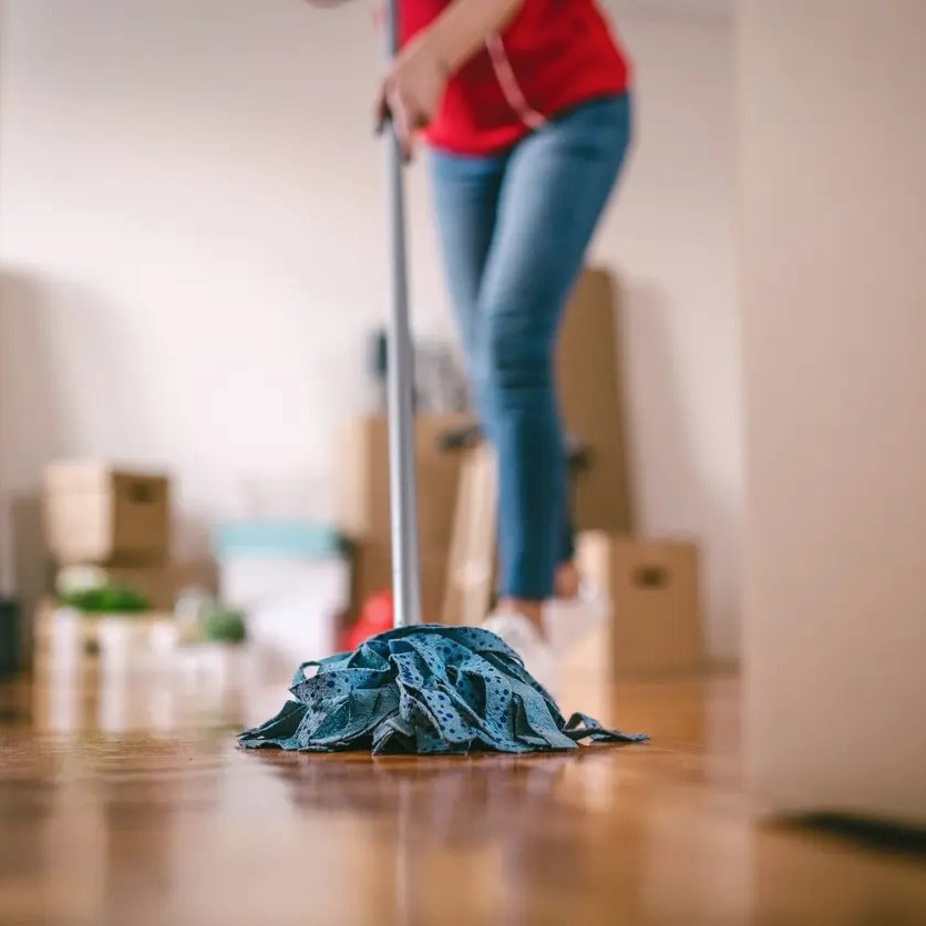A professional house cleaner mops a hardwood floor for a move while boxes sit in the background.