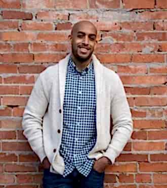 A man wearing a cream-colored cardigan and blue checkered shirt in front of a brick wall looking into the camera smiling